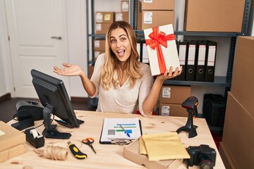 Young blonde woman working at small business ecommerce holding gift celebrating achievement with happy smile and winner expression with raised hand