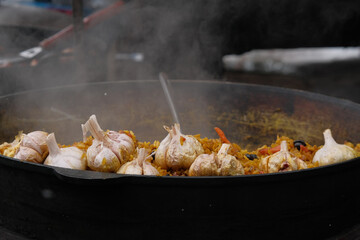 Cooking pilaf rice in a large iron pot on fire. Garlic bulb and red hot pepper