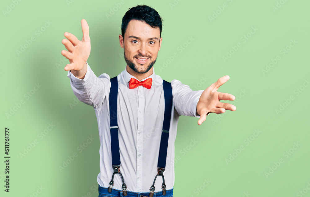 Poster Hispanic man with beard wearing hipster look with bow tie and suspenders looking at the camera smiling with open arms for hug. cheerful expression embracing happiness.