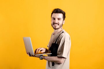Caucasian young man wearing t-shirt posing isolated over yellow background standing and using laptop pc computer smiling with a happy and cool smile on face. Showing teeth.