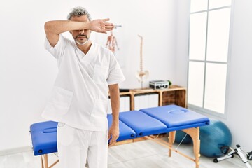 Middle age hispanic therapist man working at pain recovery clinic covering eyes with arm, looking serious and sad. sightless, hiding and rejection concept