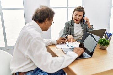 Middle age man and woman business workers using laptop and talking on the smartphone at office