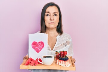 Middle age hispanic woman holding tray with breakfast food and heart draw relaxed with serious expression on face. simple and natural looking at the camera.