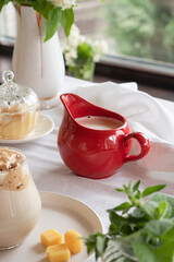 A red milk jug with fresh milk stands on a table with a white tablecloth in the middle of a green garden. Breakfast in summer.