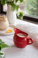 A red milk jug with fresh milk stands on a table with a white tablecloth in the middle of a green garden. Breakfast in summer.