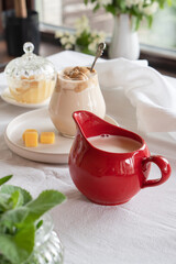 A red milk jug with fresh milk stands on a table with a white tablecloth in the middle of a green garden. Breakfast in summer.
