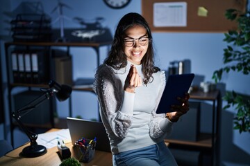 Young brazilian woman using touchpad at night working at the office doing money gesture with hands,...