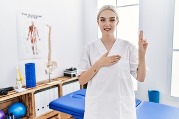 Young caucasian woman working at pain recovery clinic smiling swearing with hand on chest and...