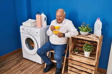 Senior man drinking coffee waiting for washing machine at laundry room