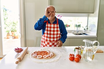 Senior man with grey hair cooking pizza at home kitchen smiling happy doing ok sign with hand on eye looking through fingers