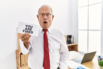 Senior business man holding you are fired banner at the office scared and amazed with open mouth for surprise, disbelief face