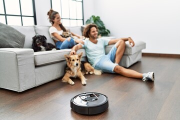 Man and woman couple sitting on sofa with dogs cleaning floor using vacuum robot at home