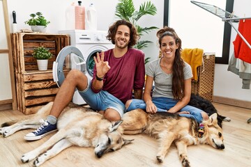 Young hispanic couple doing laundry with dogs showing and pointing up with fingers number three while smiling confident and happy.