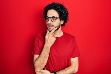 Handsome hispanic man wearing casual t shirt and glasses with hand on chin thinking about question, pensive expression. smiling with thoughtful face. doubt concept.