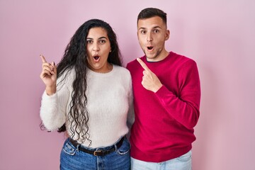 Young hispanic couple standing over pink background surprised pointing with finger to the side, open mouth amazed expression.