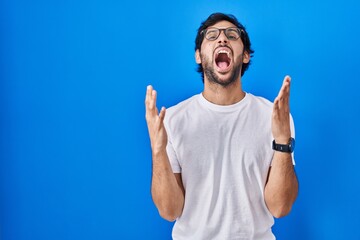 Handsome latin man standing over blue background crazy and mad shouting and yelling with aggressive expression and arms raised. frustration concept.