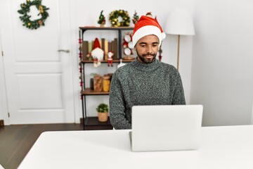 Young hispanic man with beard wearing christmas hat using laptop with serious expression on face. simple and natural looking at the camera.
