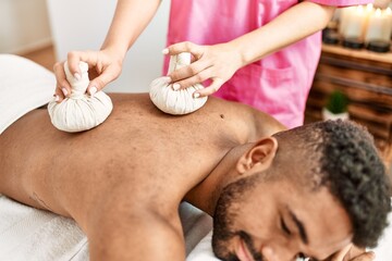 Obraz na płótnie Canvas Young african american man reciving herbal pouches thai massage at beauty center.
