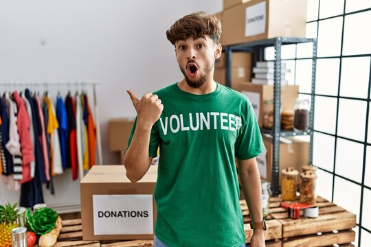 Young arab man wearing volunteer t shirt at donations stand surprised pointing with hand finger to the side, open mouth amazed expression.