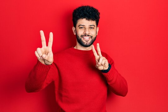 Young arab man with beard wearing casual red sweater smiling looking to the camera showing fingers doing victory sign. number two.
