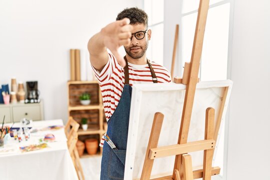 Arab young man at art studio looking unhappy and angry showing rejection and negative with thumbs down gesture. bad expression.