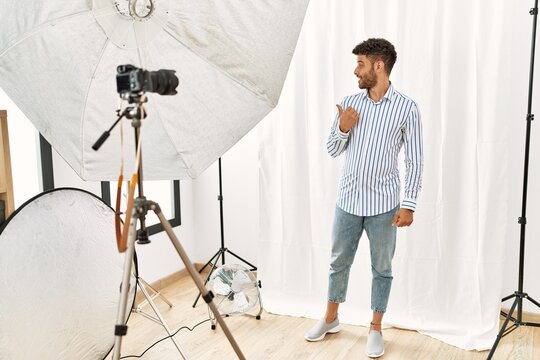 Arab young man posing as model at photography studio smiling with happy face looking and pointing to the side with thumb up.