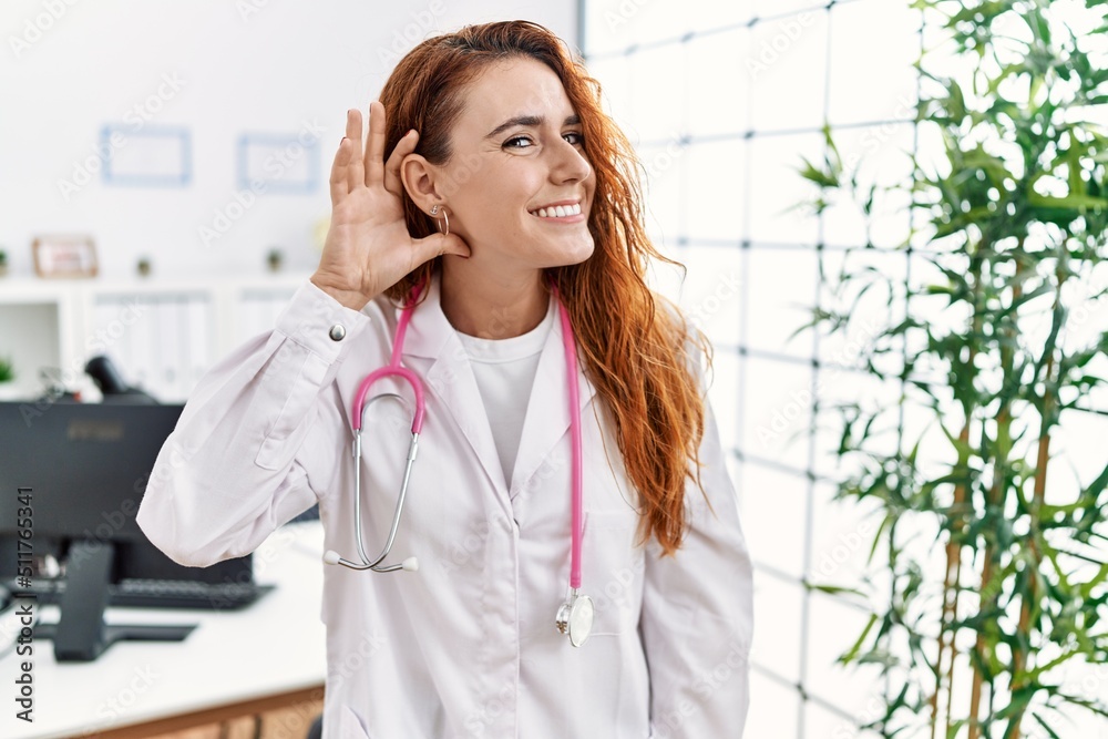 Sticker young redhead woman wearing doctor uniform and stethoscope at the clinic smiling with hand over ear 