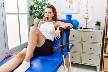 Young redhead woman lying on rehabilitation bed holding crutches surprised pointing with hand finger to the side, open mouth amazed expression.