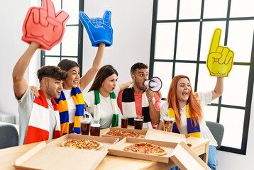 Group of young friends watching and supporting soccer match eating pizza at home.