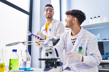 Two man scientists using microscope write on clipboard working at laboratory