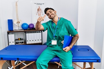 Young physiotherapist man working at pain recovery clinic approving doing positive gesture with hand, thumbs up smiling and happy for success. winner gesture.