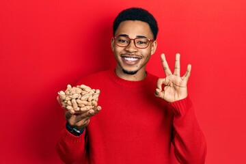 Young african american man holding peanuts doing ok sign with fingers, smiling friendly gesturing excellent symbol