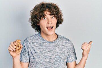 Handsome young man eating protein bar as healthy energy snack pointing thumb up to the side smiling...