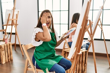 Young hispanic artist women painting on canvas at art studio covering one eye with hand, confident smile on face and surprise emotion.