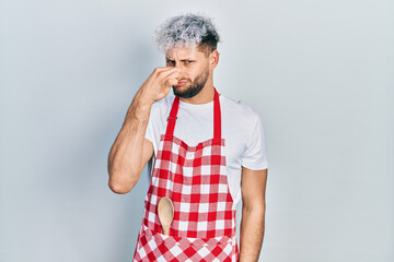 Young hispanic man with modern dyed hair wearing apron smelling something stinky and disgusting,...
