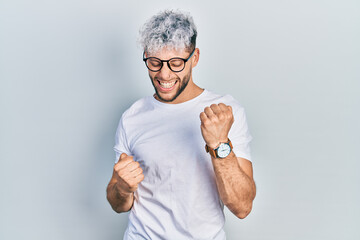 Young hispanic man with modern dyed hair wearing white t shirt and glasses celebrating surprised...