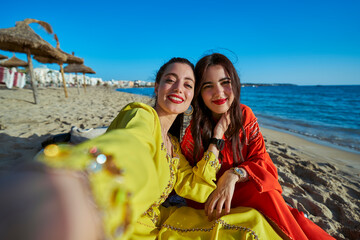 Two Arab women, photo taken in selfie mode on the beach sitting with typical Moroccan clothes....