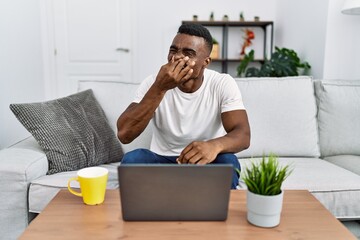 Young african man using laptop at home smelling something stinky and disgusting, intolerable smell,...