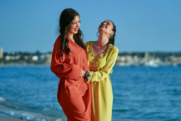 Two Arab women strolling along the beach in typical Moroccan clothing. Enjoying the summer, having a great time. Horizontal photo