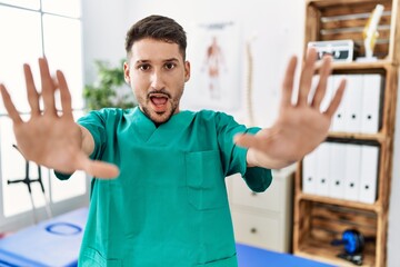 Young physiotherapist man working at pain recovery clinic doing stop gesture with hands palms, angry and frustration expression