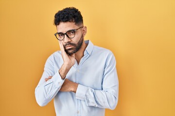 Hispanic man with beard standing over yellow background thinking looking tired and bored with...