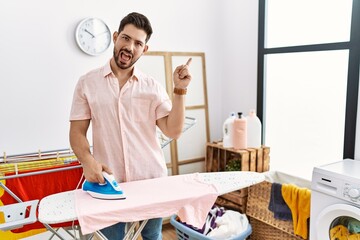 Young man with beard ironing clothes at home with a big smile on face, pointing with hand finger to the side looking at the camera.