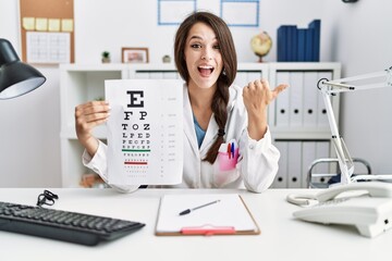 Young doctor woman holding eyesight test pointing thumb up to the side smiling happy with open mouth