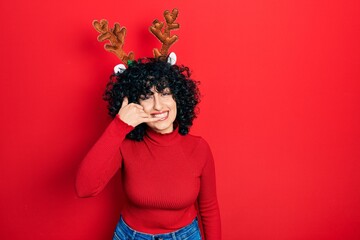 Young middle east woman wearing cute christmas reindeer horns smiling doing phone gesture with hand...