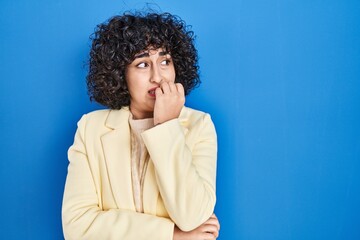 Fototapeta na wymiar Young brunette woman with curly hair standing over blue background looking stressed and nervous with hands on mouth biting nails. anxiety problem.
