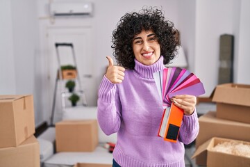 Young brunette woman with curly hair choosing color of new house wall smiling happy and positive, thumb up doing excellent and approval sign