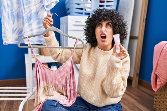 Young brunette woman with curly hair dyeing tye die dress with pink color angry and mad screaming frustrated and furious, shouting with anger looking up.