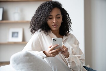 Smiling young mixed race woman sit on sofa at home using cellphone texting chatting with friend