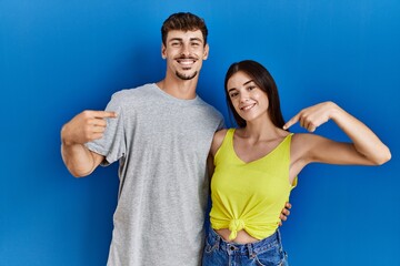 Young hispanic couple standing together over blue background looking confident with smile on face, pointing oneself with fingers proud and happy.