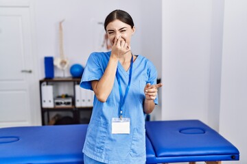 Young hispanic woman wearing physiotherapist uniform standing at clinic smelling something stinky and disgusting, intolerable smell, holding breath with fingers on nose. bad smell - obrazy, fototapety, plakaty